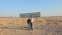Pareja posando junto al letrero del Trópico de Capricornio en un paisaje árido  